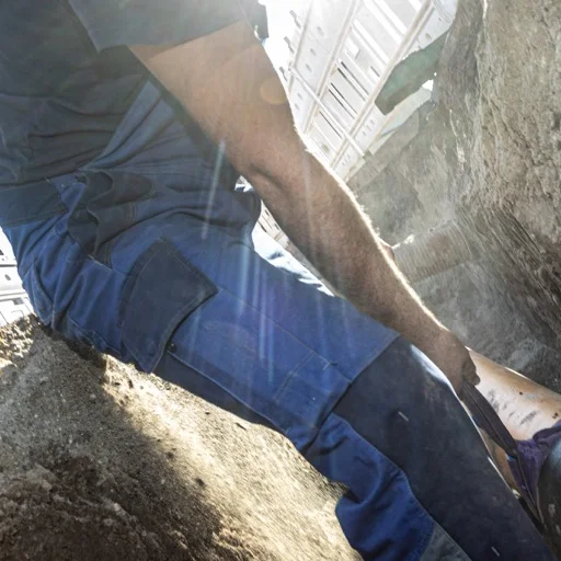 Ein Mann in einem blauen Arbeitsanzug platziert bei strahlendem Sonnenschein und blauem Himmel sorgfältig ein Rohr in einem vorbereiteten Baugraben.